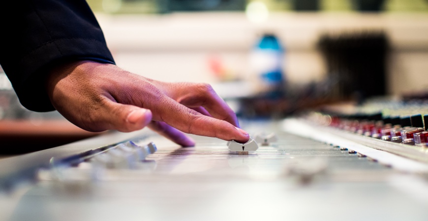 Wedding DJ Miami A person adjusts sliders on a sound mixing console in a recording studio. The close-up shows their hand on the panel, with buttons and knobs blurred in the background.