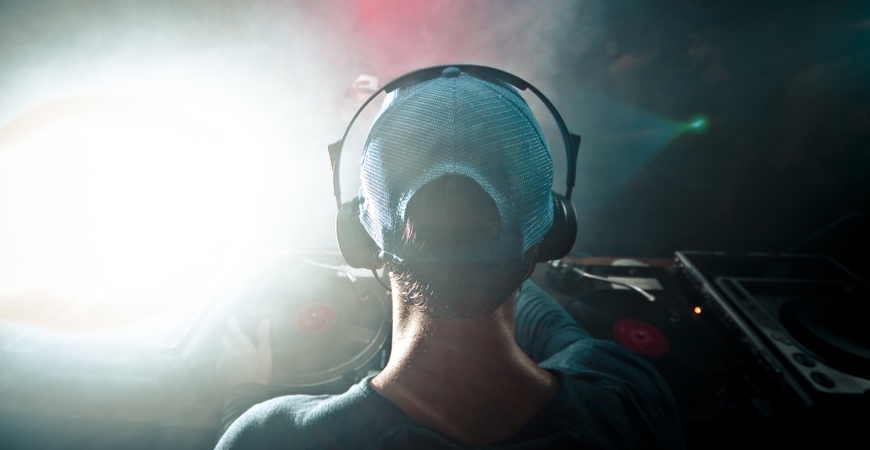 Wedding DJ Miami A DJ wearing a blue cap and headphones stands at a turntable, surrounded by dim lighting and smoke, with lights creating a dramatic effect in the background.