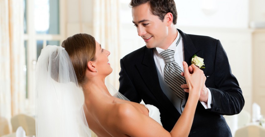 Wedding DJ Miami A bride and groom smile at each other while dancing. The bride is wearing a white wedding dress and veil, and the groom is in a black suit with a silver tie and boutonniere. The setting appears to be a bright, elegantly decorated indoor venue.