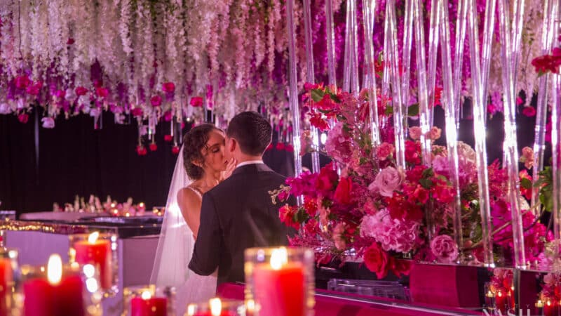 A bride and groom under a ceiling treatment full of hanging flowers with beautiful pink lighting